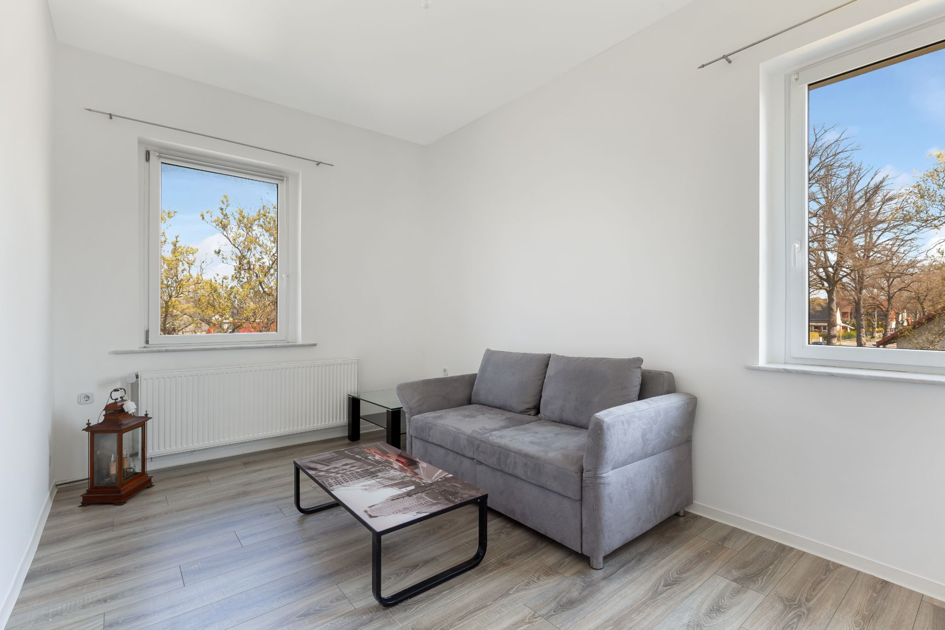Helles Wohnzimmer mit grauem Sofa, Couchtisch, zwei Fenstern und Laterne auf Holzboden.
