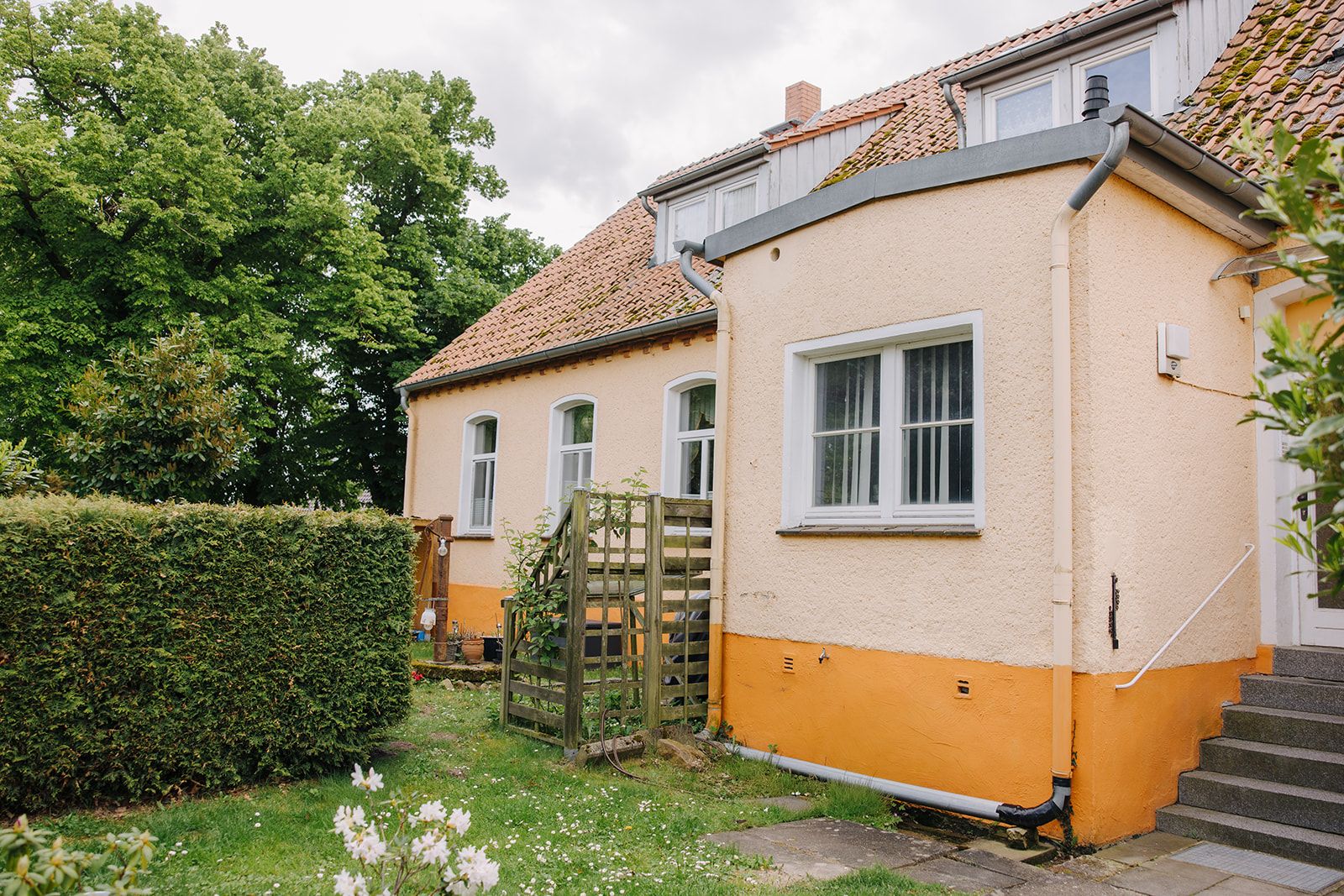 Gelbes Wohnhaus mit roten Dachziegeln, Garten, Hecke und Blumenecke im Vordergrund.