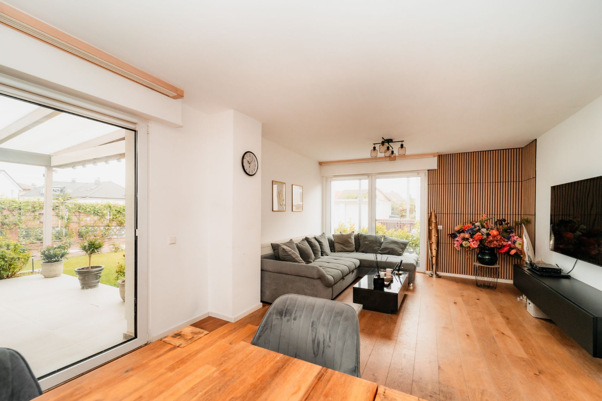 Helles Wohnzimmer mit Holzfußboden, grauem Sofa, Wandfernseher und Zugang zur Terrasse mit Pflanzen.