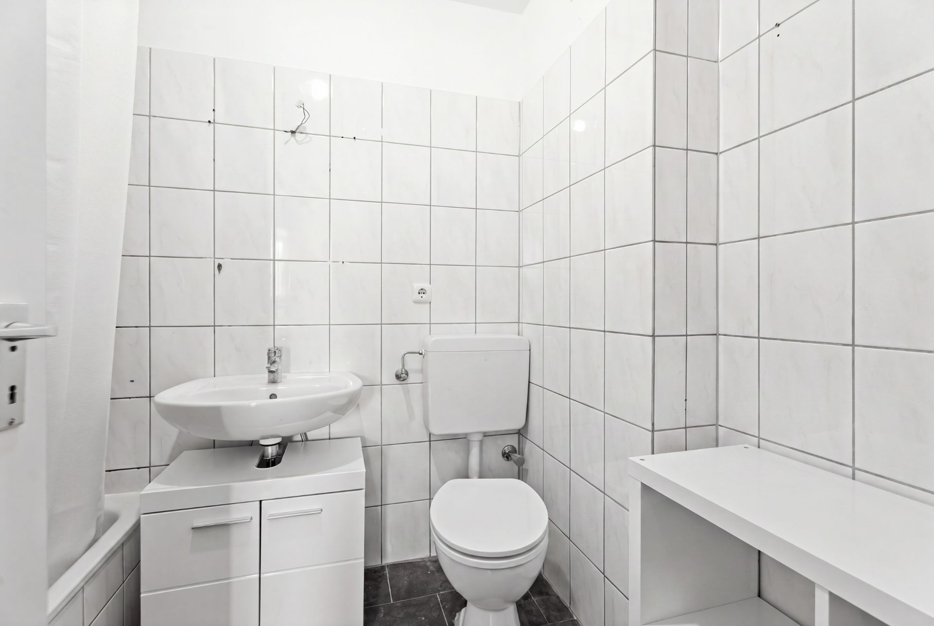 Kleines Bad mit weiß gefliesten Wänden, Waschbecken, Toilette und Regal.