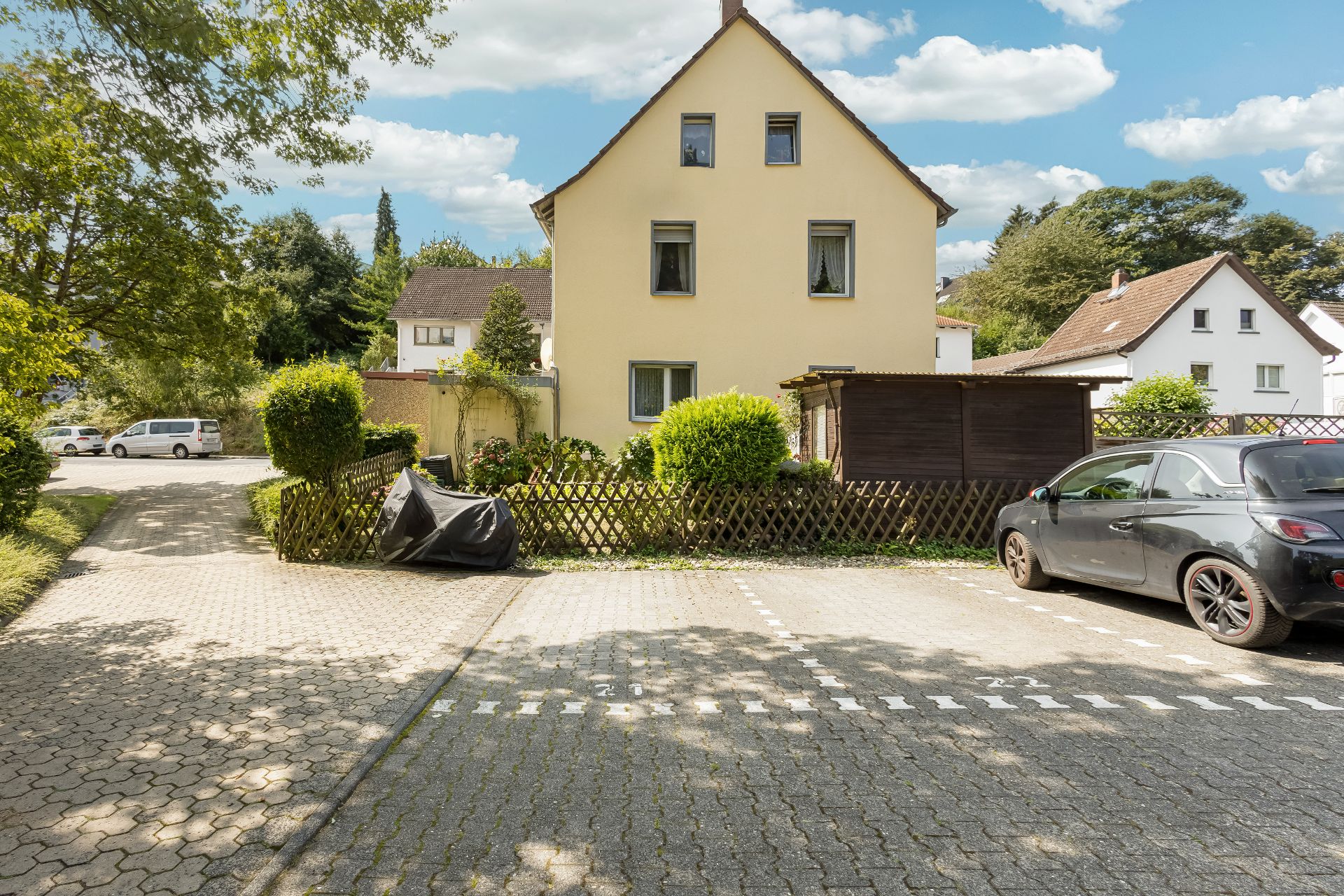 Zweistöckiges Wohnhaus mit Garten, Auto auf Parkplatz und Bäumen im Hintergrund.