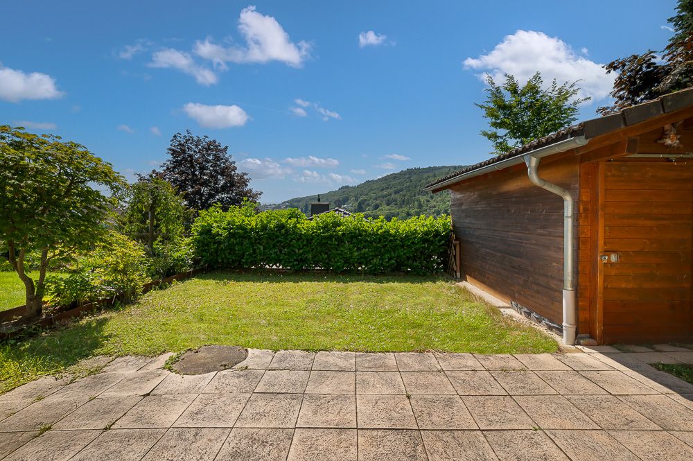 Gepflasterte Terrasse mit Rasenfläche und Holzhütte im Garten, umgeben von Büschen und Bäumen.