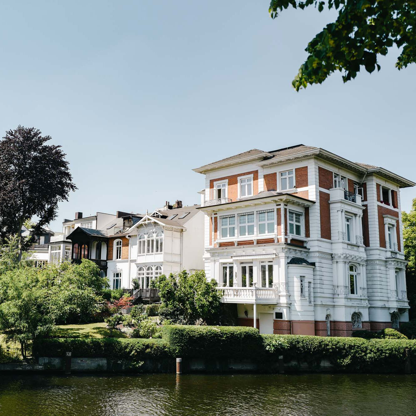 Großes Wohnhaus mit Garten direkt an einem Fluss, daneben weitere Häuser und viel Grün.