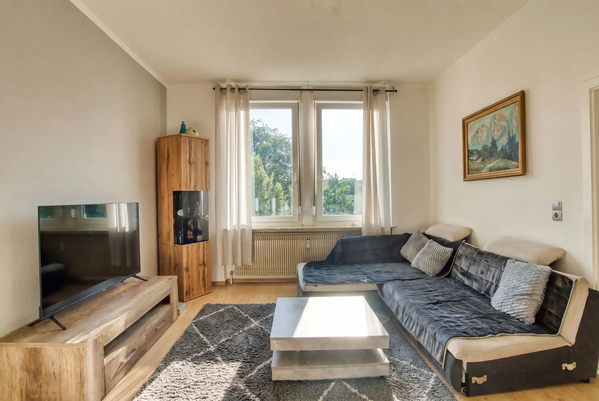 Wohnzimmer mit grauem Sofa, Couchtisch, Teppich, TV und großem Fenster mit Vorhängen.
