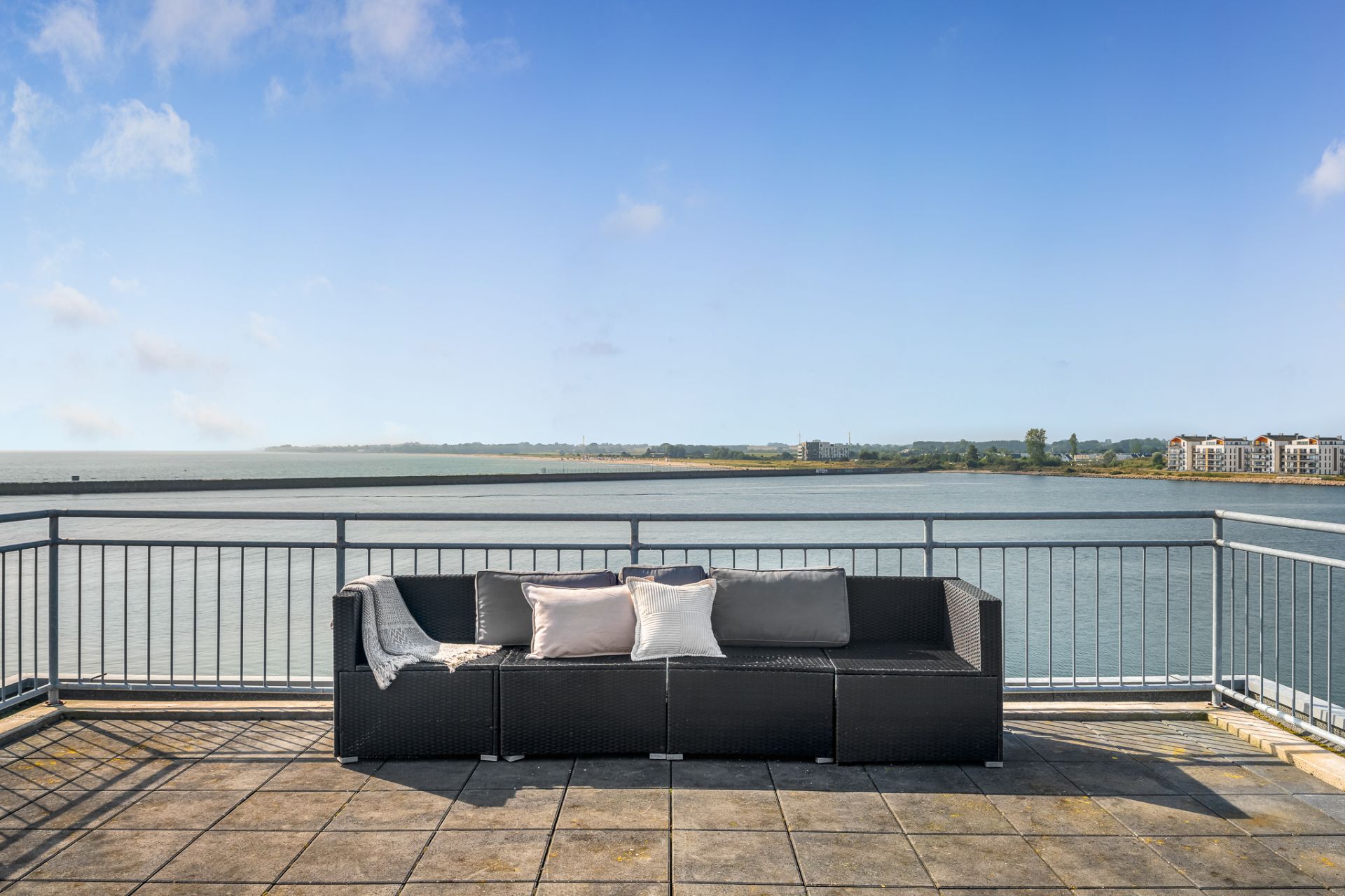 Terrasse mit Sofa und Kissen, Blick auf Wasser und entfernte Gebäude.