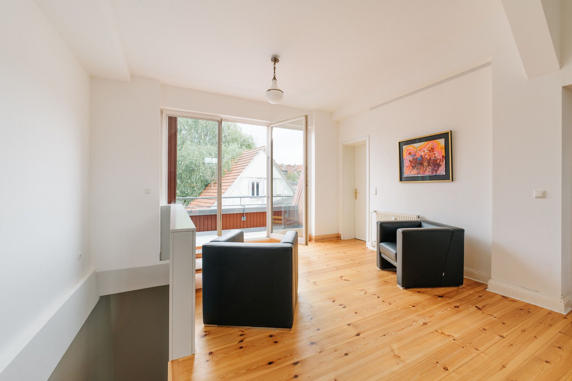 Heller Raum mit Holzboden, zwei schwarzen Sesseln, Bild an der Wand und großen Fenstern.