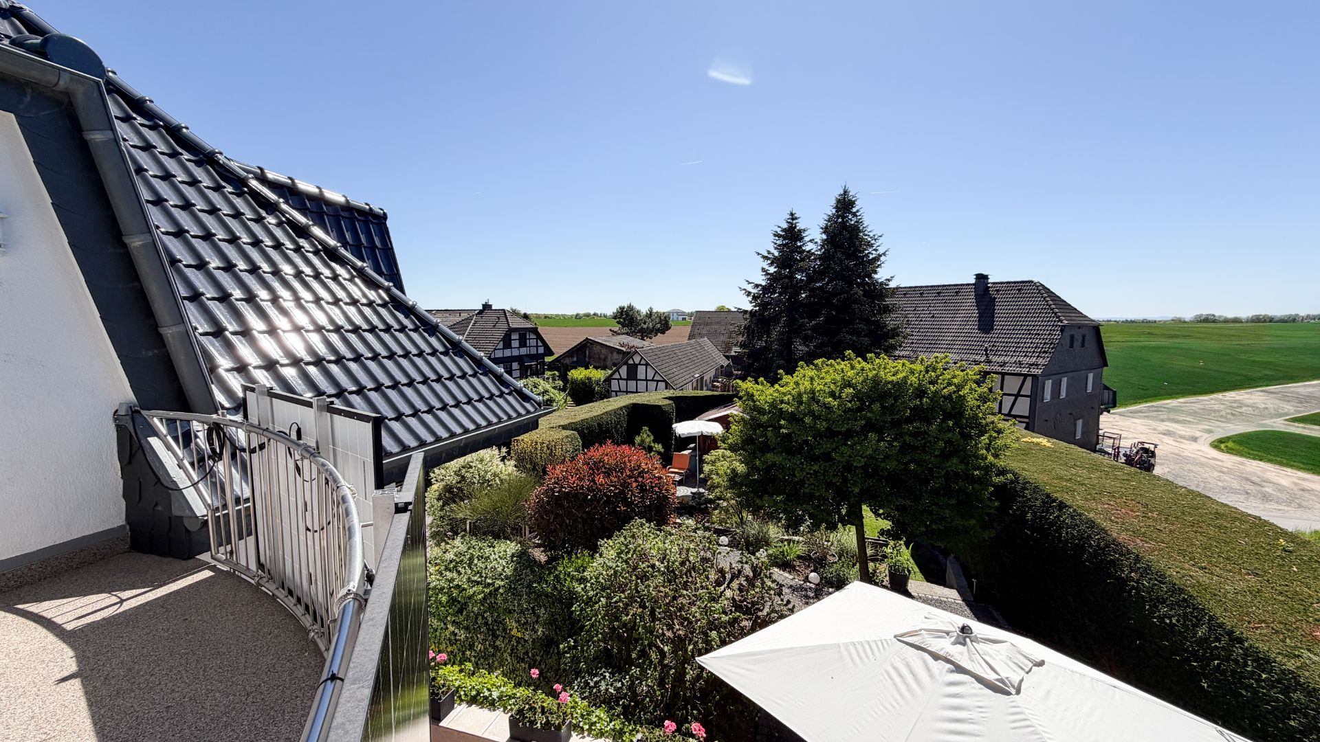 Blick vom Balkon auf einen Garten, Nachbarhäuser und weite grüne Felder bei klarem Himmel.