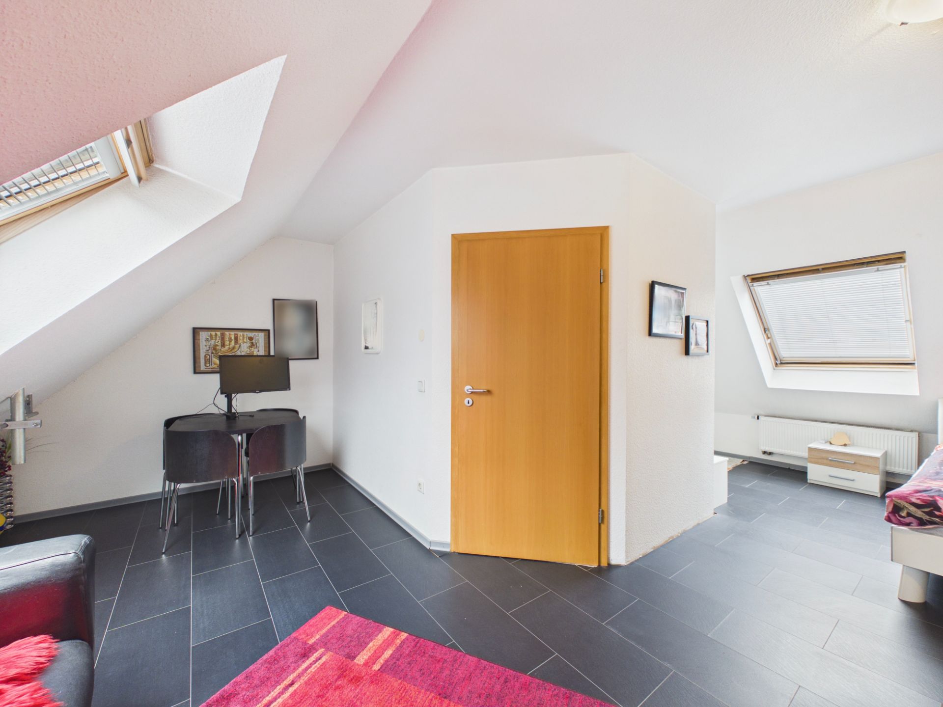 Helles Zimmer mit Dachfenstern, Holztür, Schreibtisch und Stuhl, schwarzer Fliesenboden.