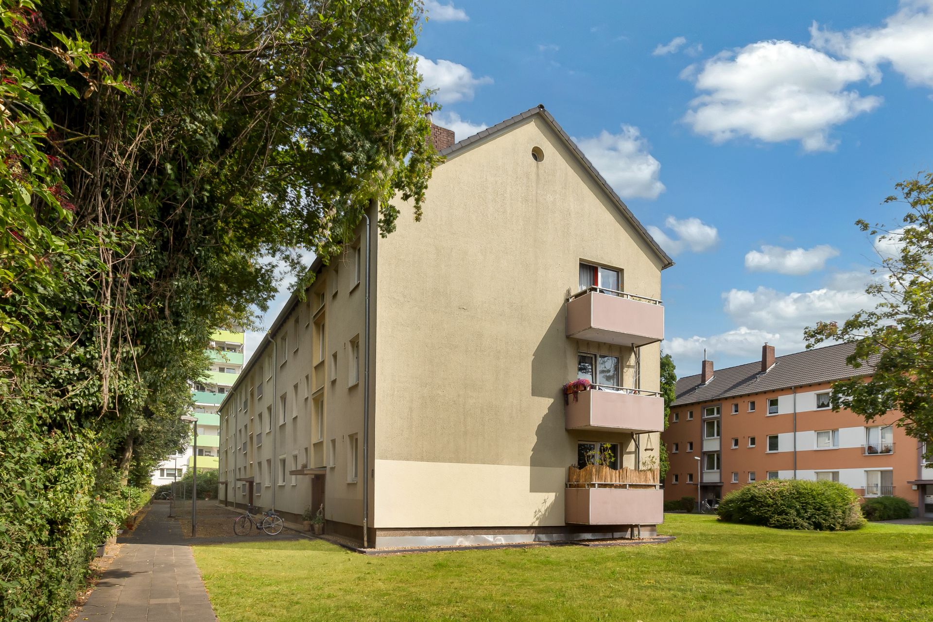 Mehrfamilienhaus mit Balkonen und Rasenfläche davor, seitlich Bäume und Gehweg.