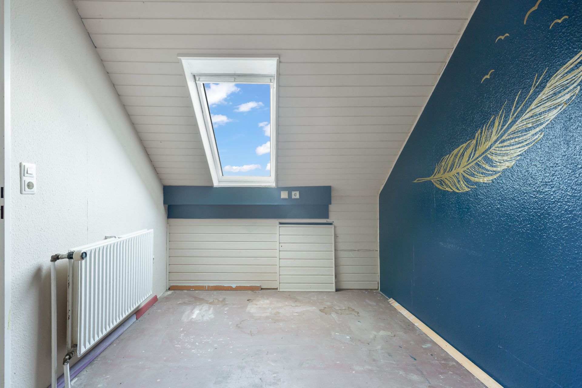 Kleines Zimmer mit Dachfenster, blauem Wandbild mit Feder und Heizkörper.