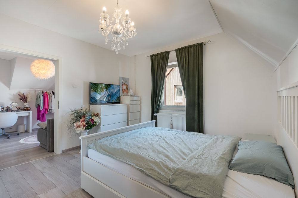 Helles Schlafzimmer mit Doppelbett, Kronleuchter, Kommode und Fenster mit dunklen Vorhängen.