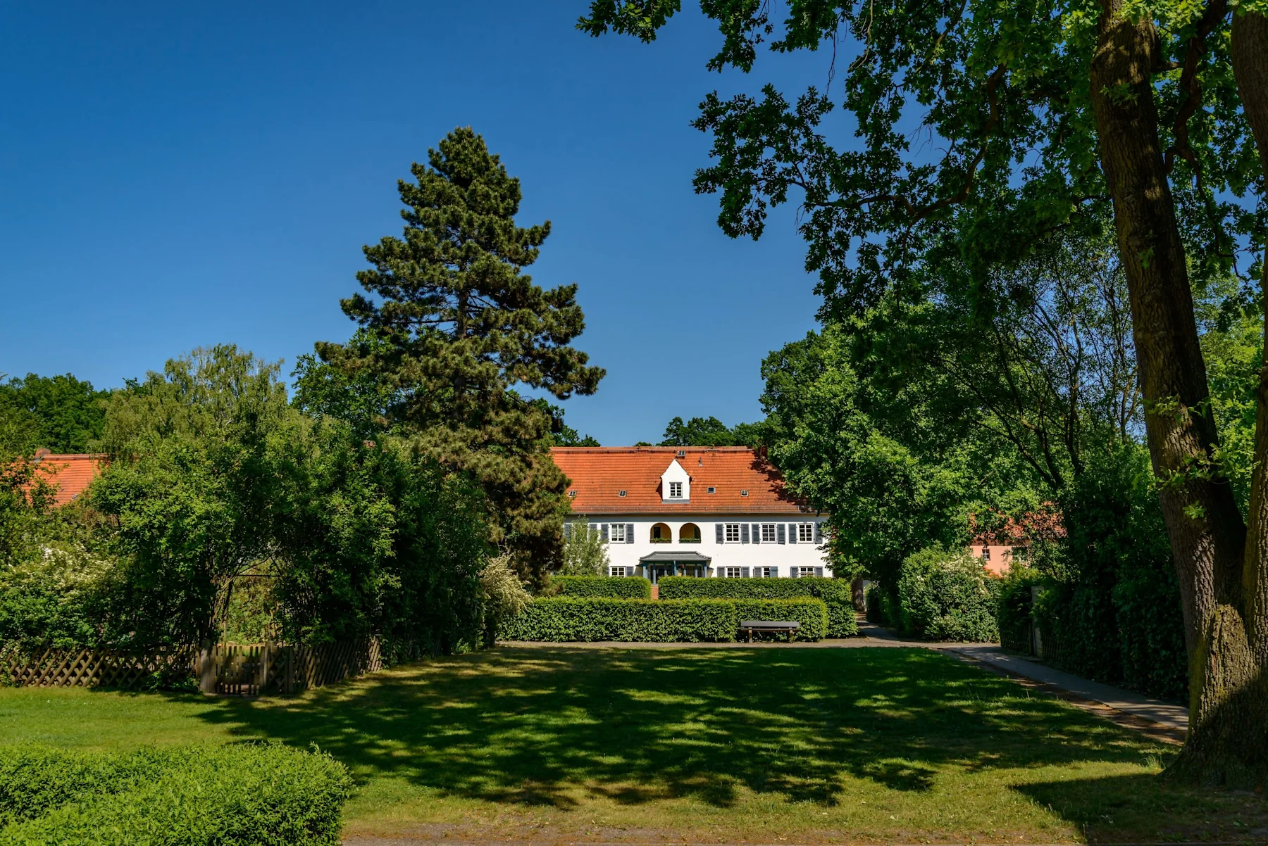 Weißes Wohnhaus mit rotem Dach, großer Garten und viele Bäume bei sonnigem Wetter.