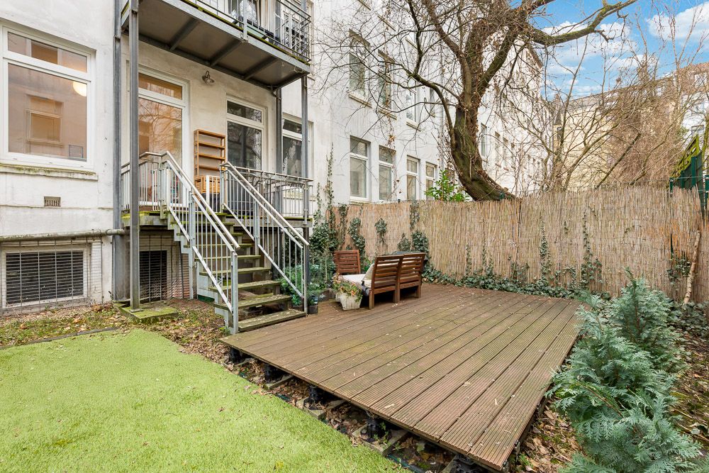 Holzterrasse mit Sitzmöbeln im Garten neben einem Wohngebäude, mit Treppe und grünem Rasen.