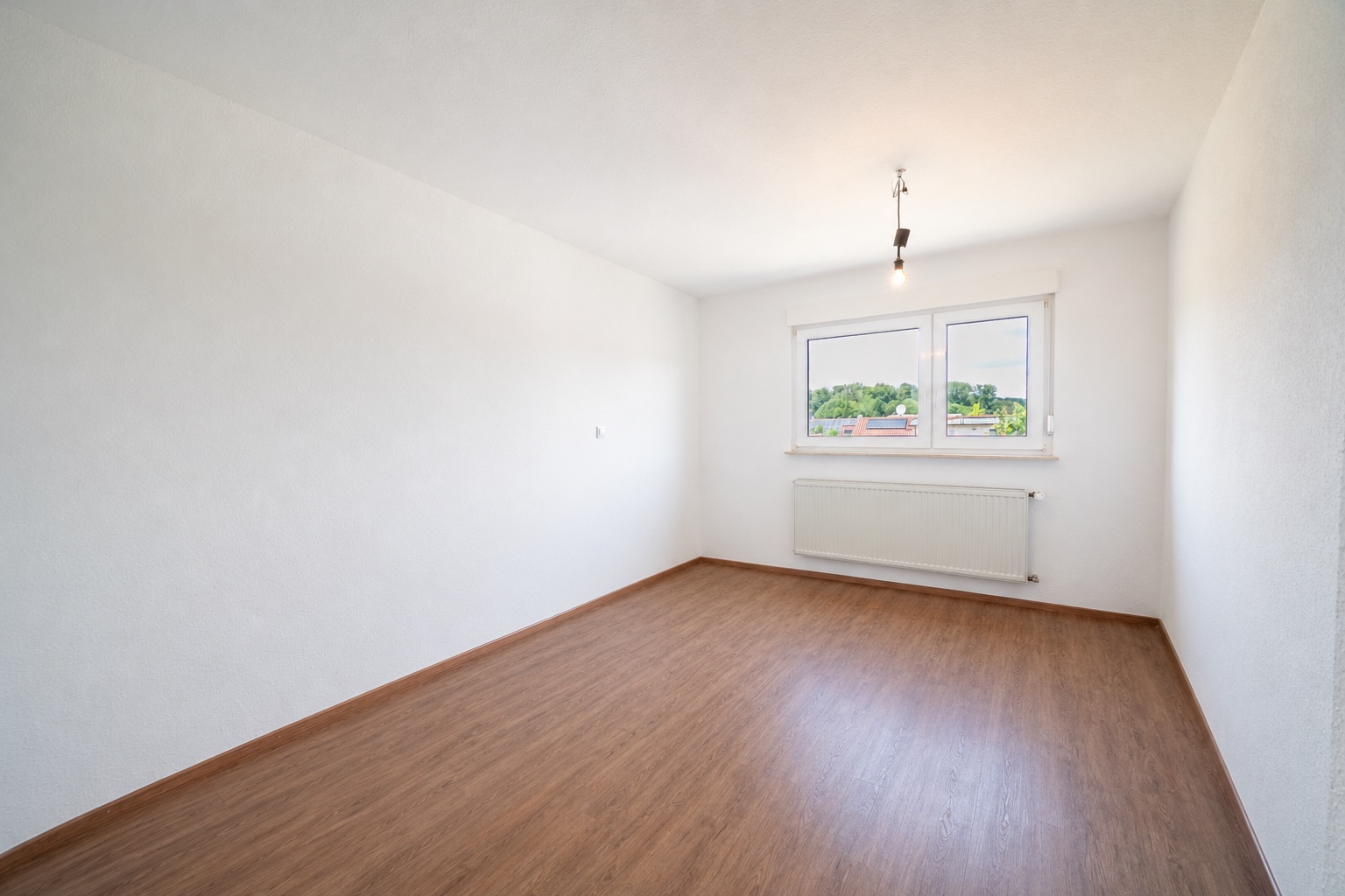 Leeres Zimmer mit weißem Wänden, Holzboden, Fenster und Heizkörper unter dem Fenster.