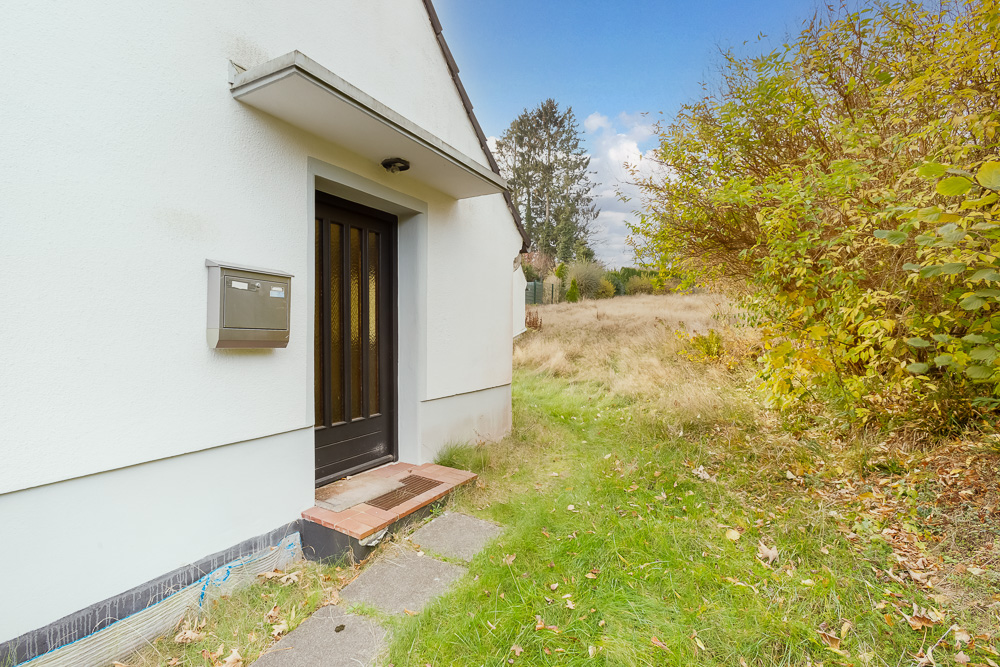 Seiteneingang eines Wohnhauses mit Briefkasten und Garten mit Büschen und Wiese.