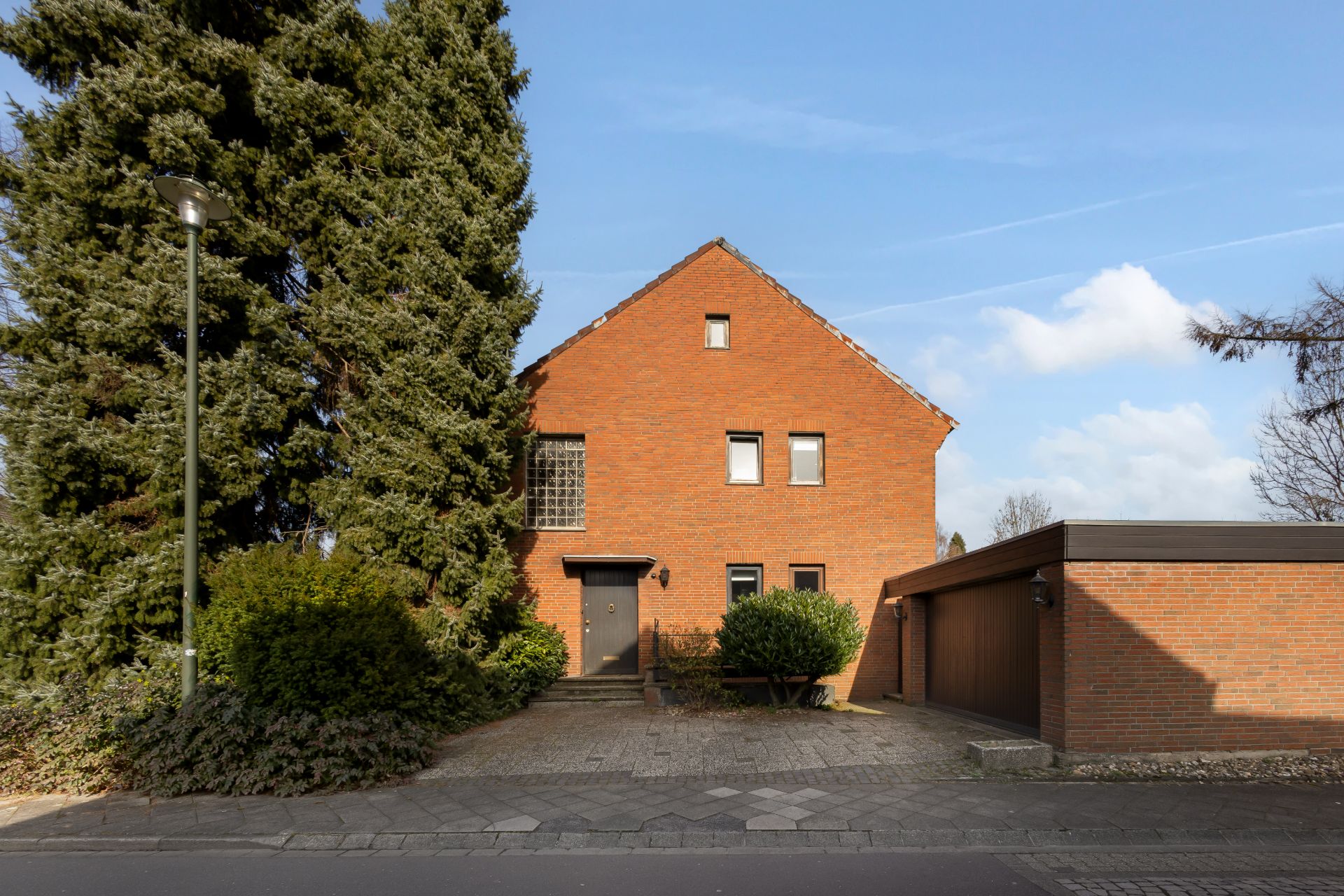 Backsteinhaus mit Satteldach, großer Baum links, Einfahrt und Garage rechts.