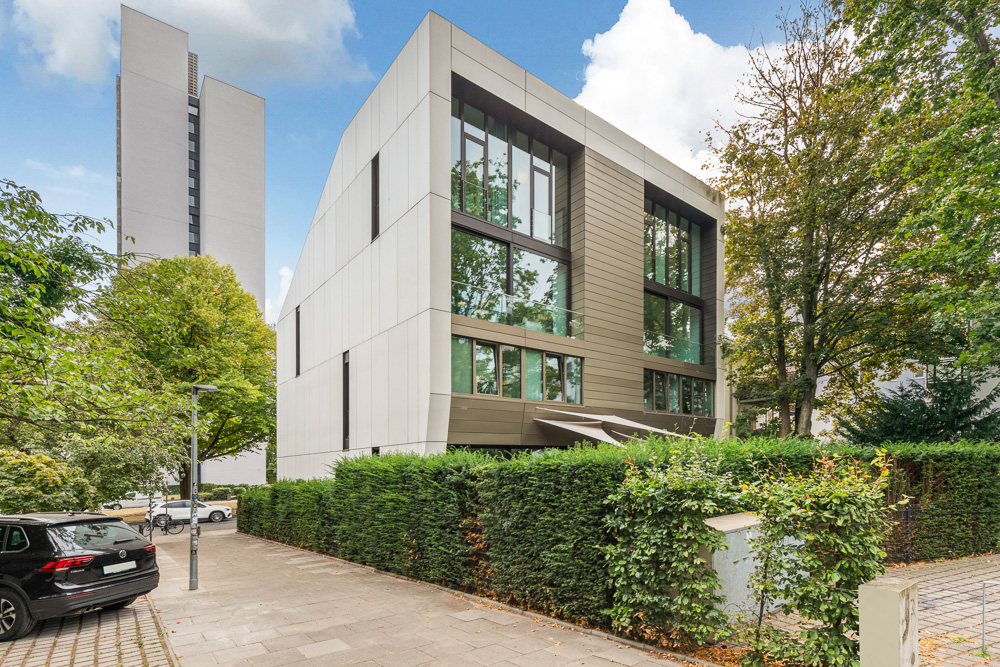 Modernes Wohngebäude mit großen Fenstern und umgebender Hecke an einer Straße.