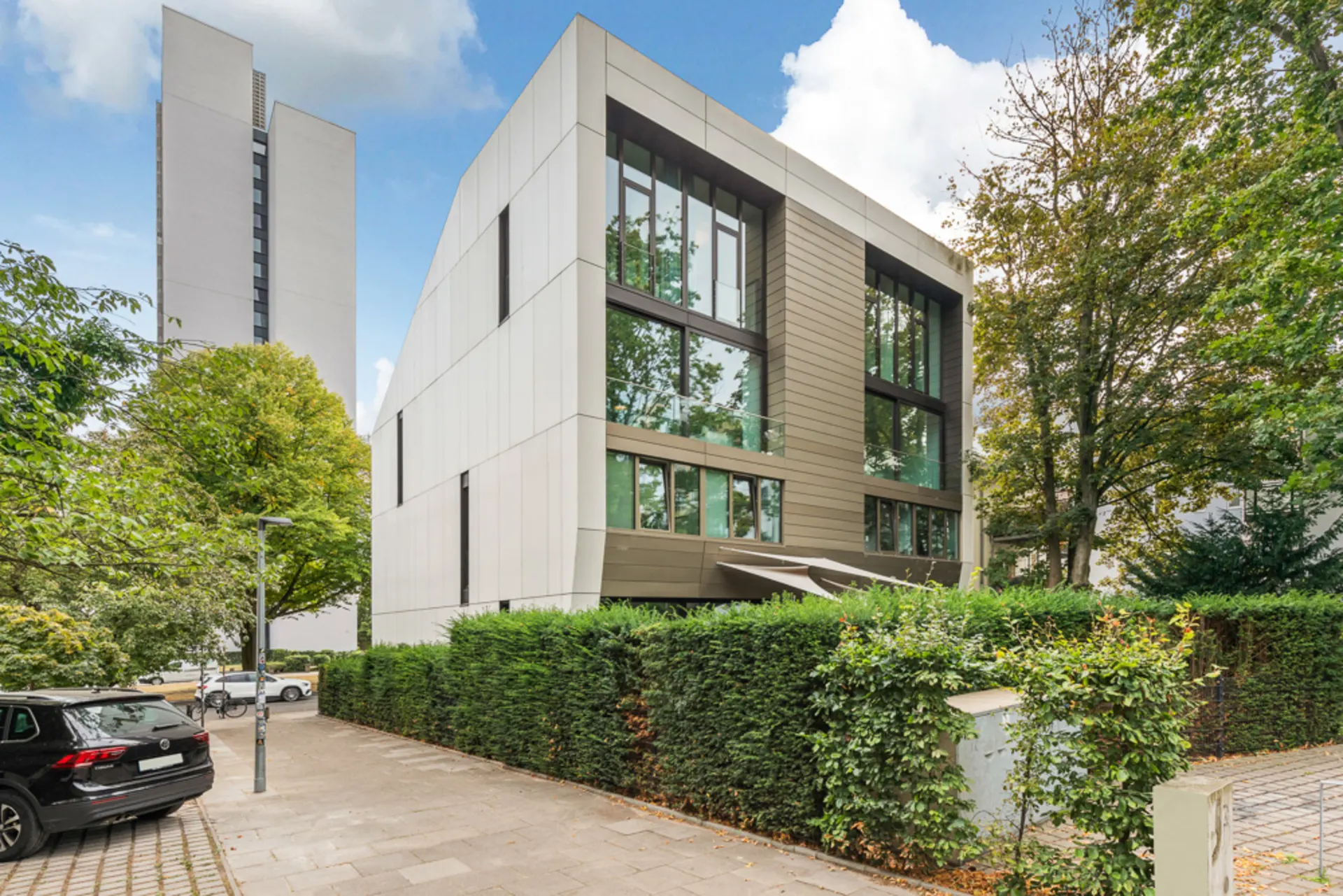Modernes Wohngebäude mit großen Fenstern und umgebender Hecke an einer Straße.