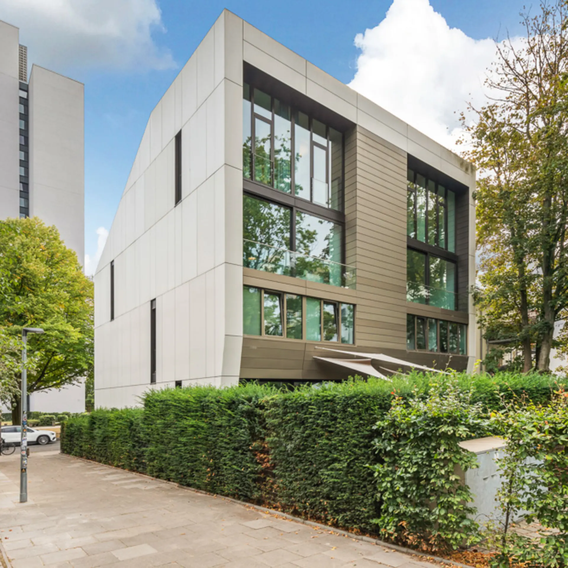 Modernes Wohngebäude mit großen Fenstern und umgebender Hecke an einer Straße.