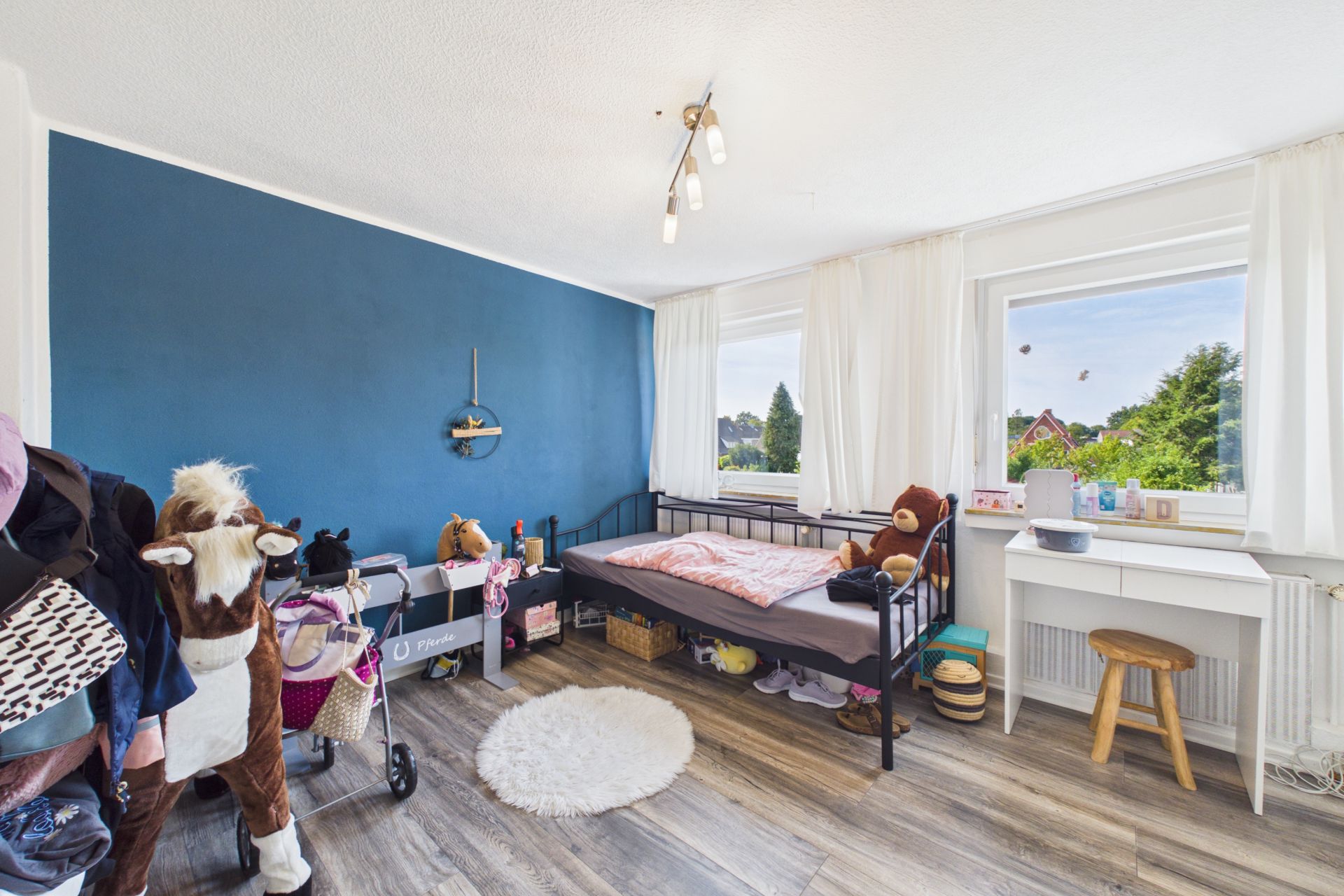 Helles Kinderzimmer mit blau gestrichener Wand, Einzelbett, Spielzeugpferd und Schreibtisch am Fenster.