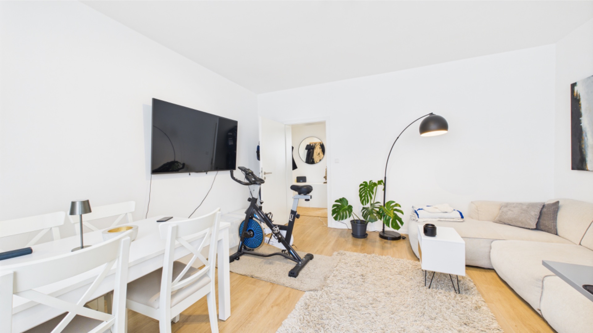 Helles Wohnzimmer mit Sofa, Esstisch, Fernseher, Hometrainer und Pflanzen auf Holzfußboden.
