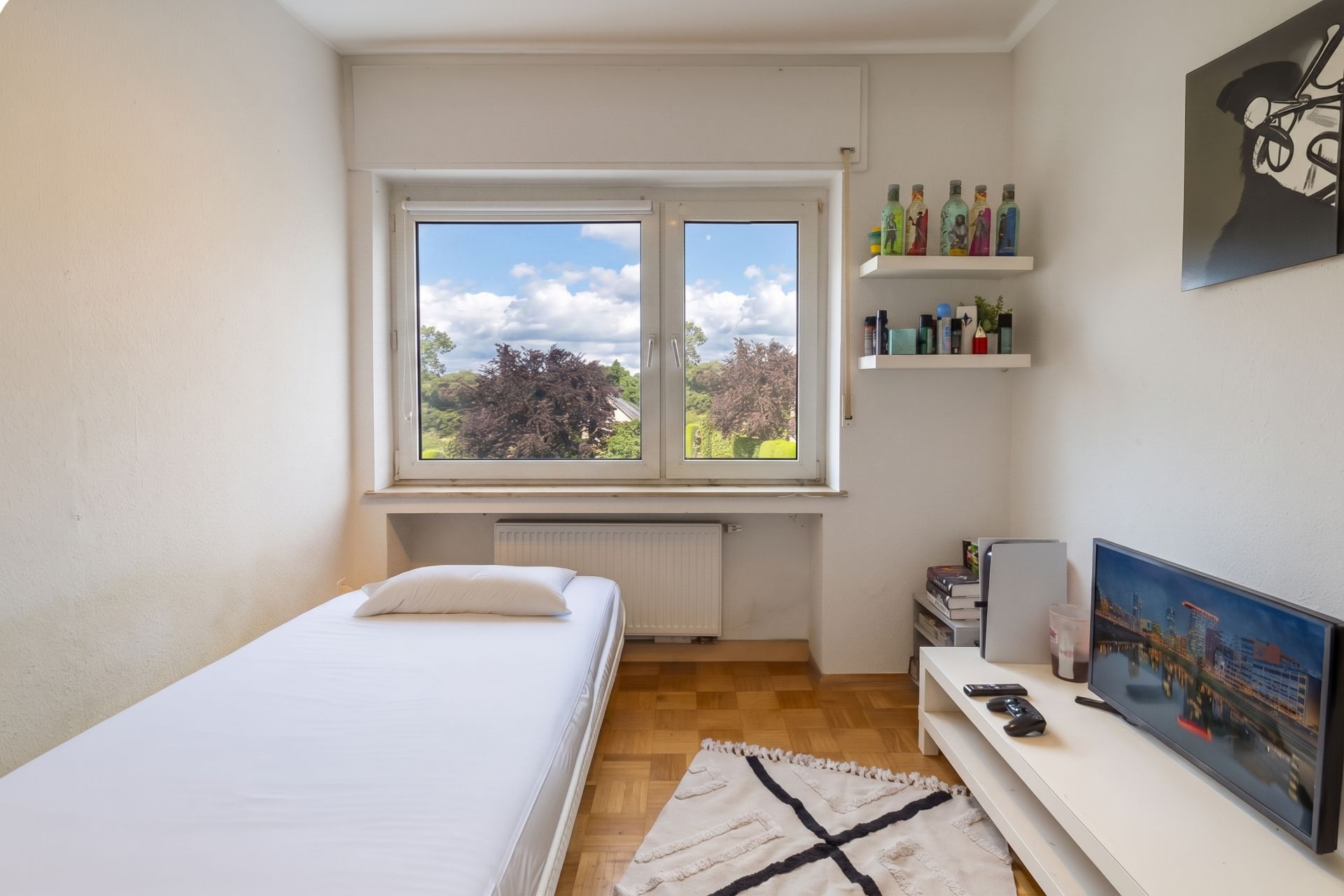 Kleines Schlafzimmer mit Einzelbett, Fenster, Regal, TV und Teppich auf Parkettboden.