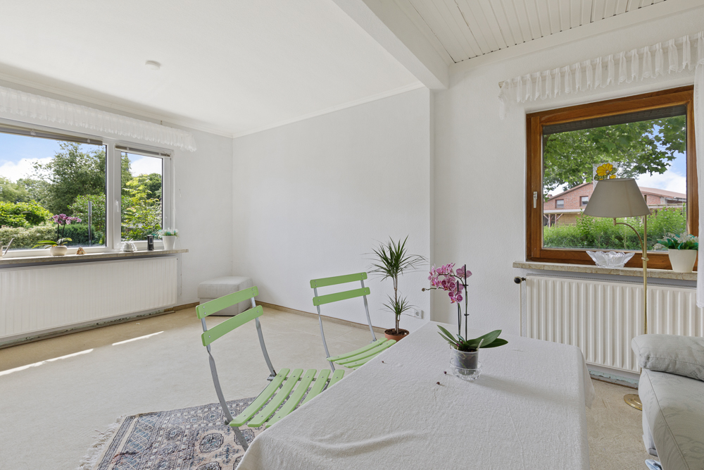Helles Wohnzimmer mit zwei grünen Stühlen, Tisch, Pflanzen und großen Fenstern mit Blick ins Grüne.