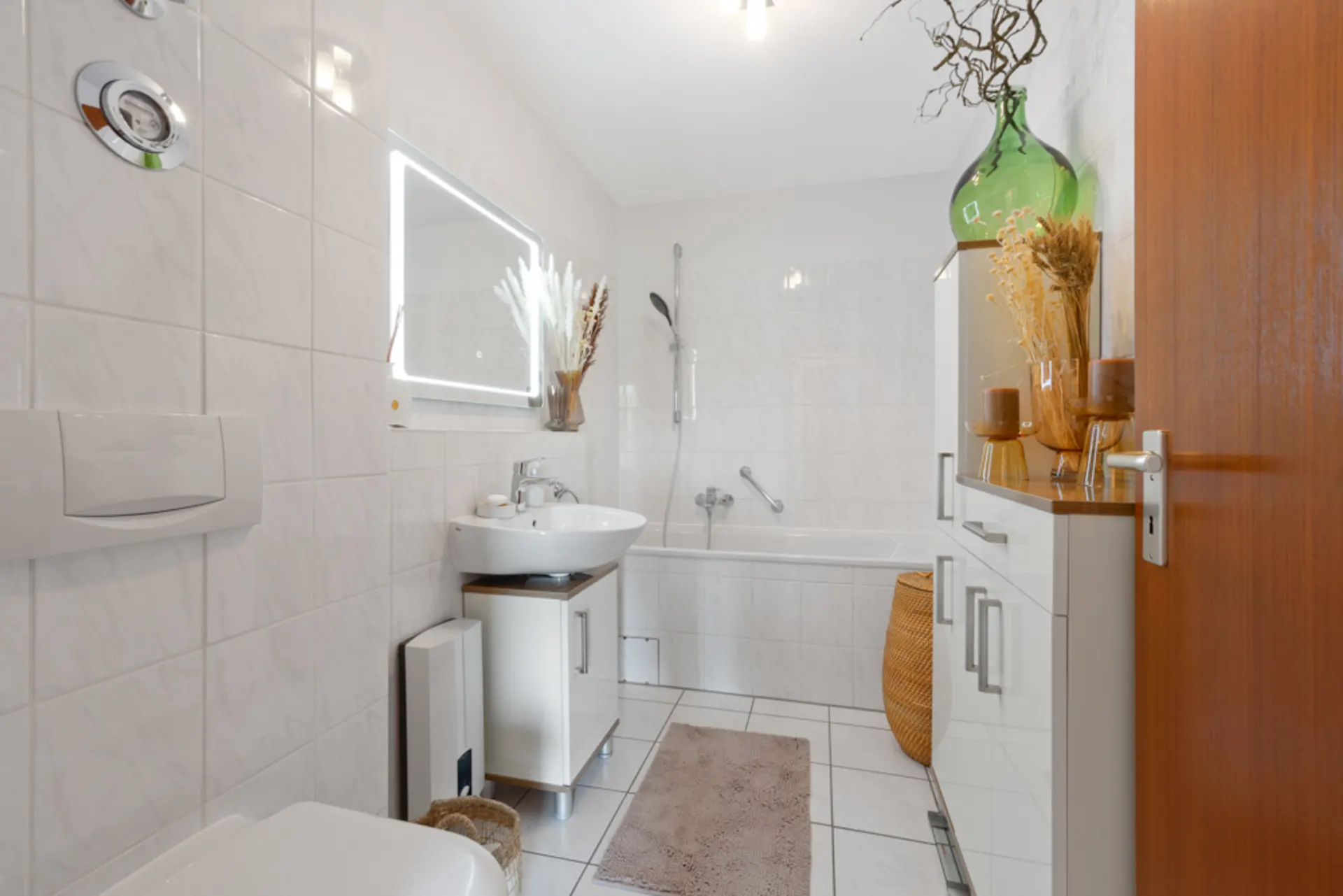 Helles Bad mit Badewanne, Waschbecken, Spiegel und weißen Fliesen, dekoriert mit Vasen und Trockenblumen.