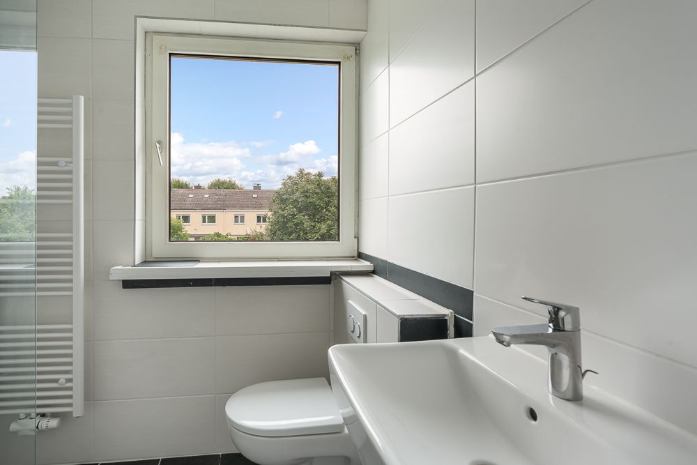 Helles Badezimmer mit weißem Waschbecken, Toilette und Fenster mit Blick nach draußen.