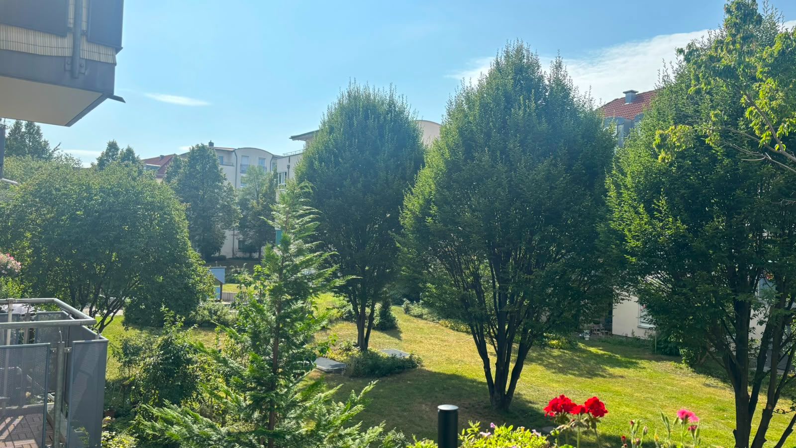 Grüner Garten mit mehreren Bäumen, Blumenbeet und angrenzenden Wohngebäuden bei sonnigem Wetter.