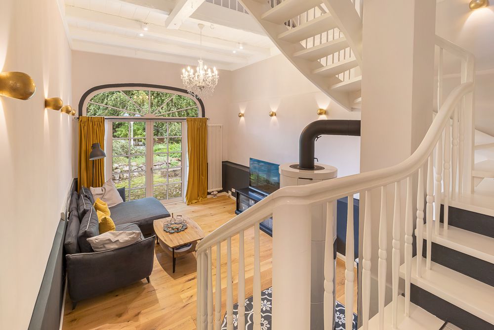 Heller Wohnraum mit Sofa, Gelbvorhängen, Kamin, TV und Treppe, große Fenster mit Blick in den Garten.
