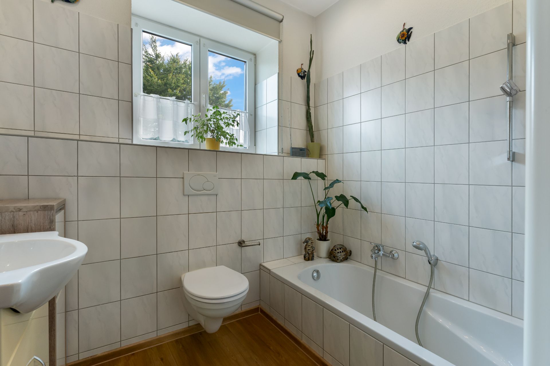 Helles Badezimmer mit Badewanne, wandhängender Toilette, Waschbecken und Fenster mit Pflanzen.