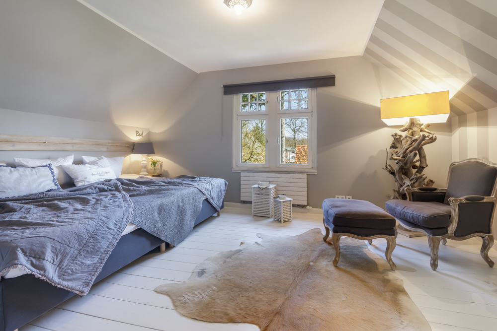 Helles Schlafzimmer mit großem Bett, Sessel, Hocker, Fenster und Tierfell auf dem Boden.