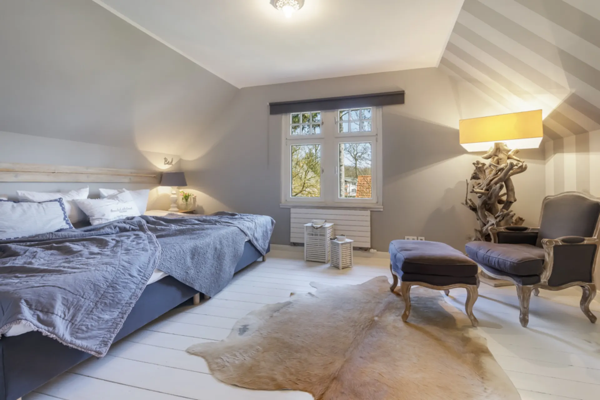 Helles Schlafzimmer mit großem Bett, Sessel, Hocker, Fenster und Tierfell auf dem Boden.