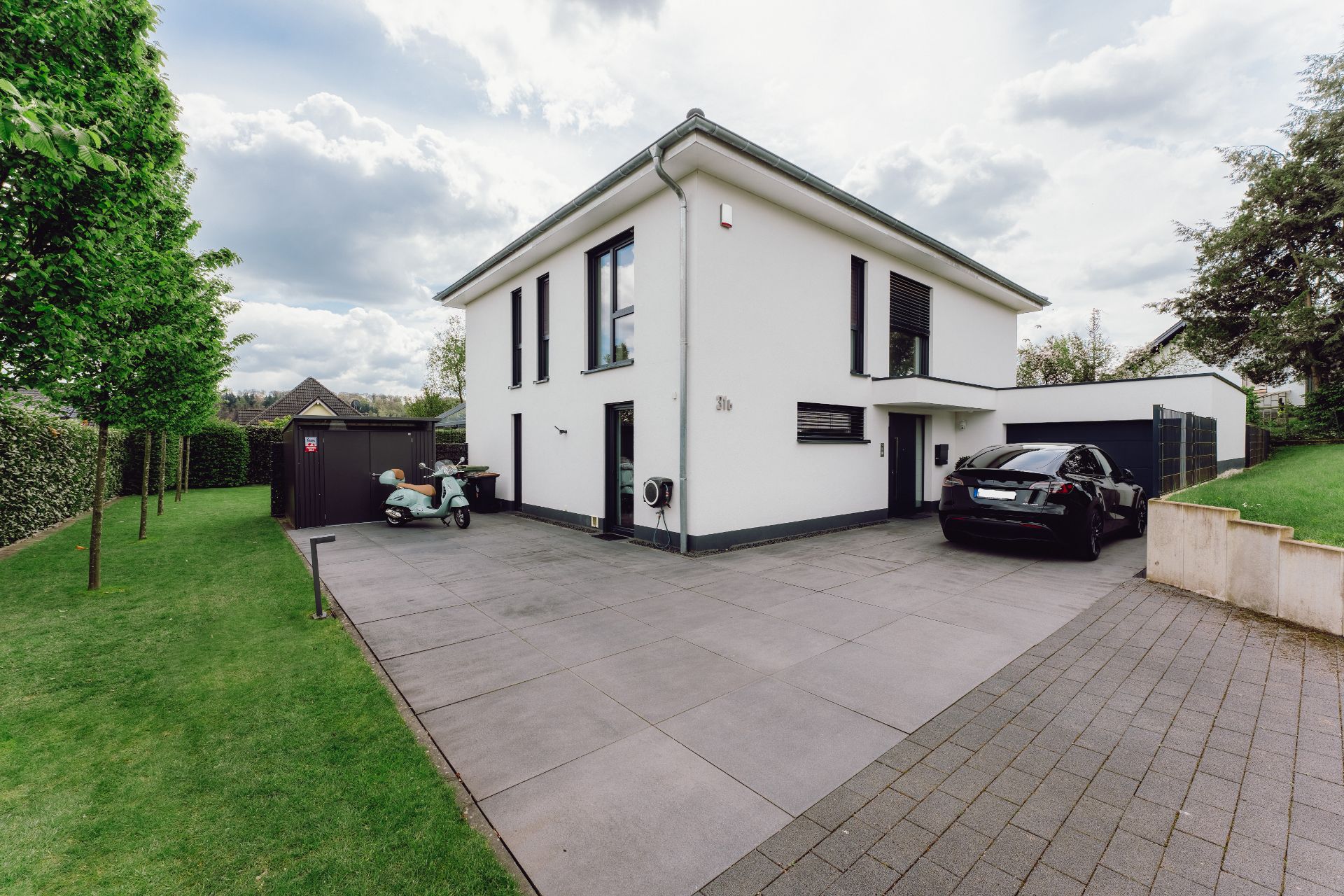 Modernes weißes Wohnhaus mit gepflasterter Einfahrt, schwarzem Auto und Roller, umgeben von Rasen.