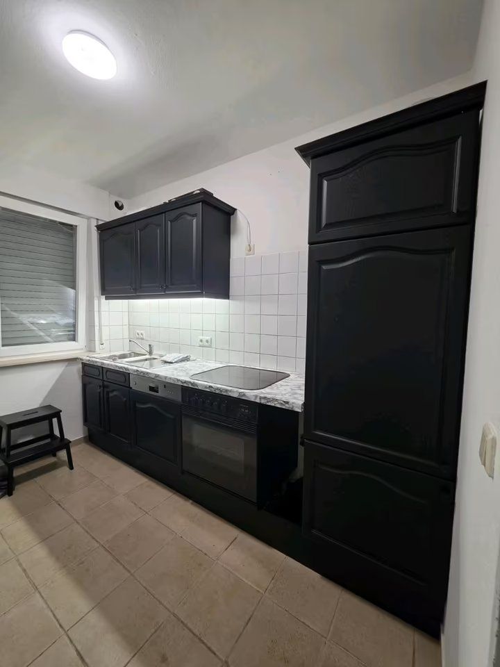 Küche mit schwarzen Schränken, Herd, Spüle, Fenster und kleinem Hocker auf beigem Fliesenboden.