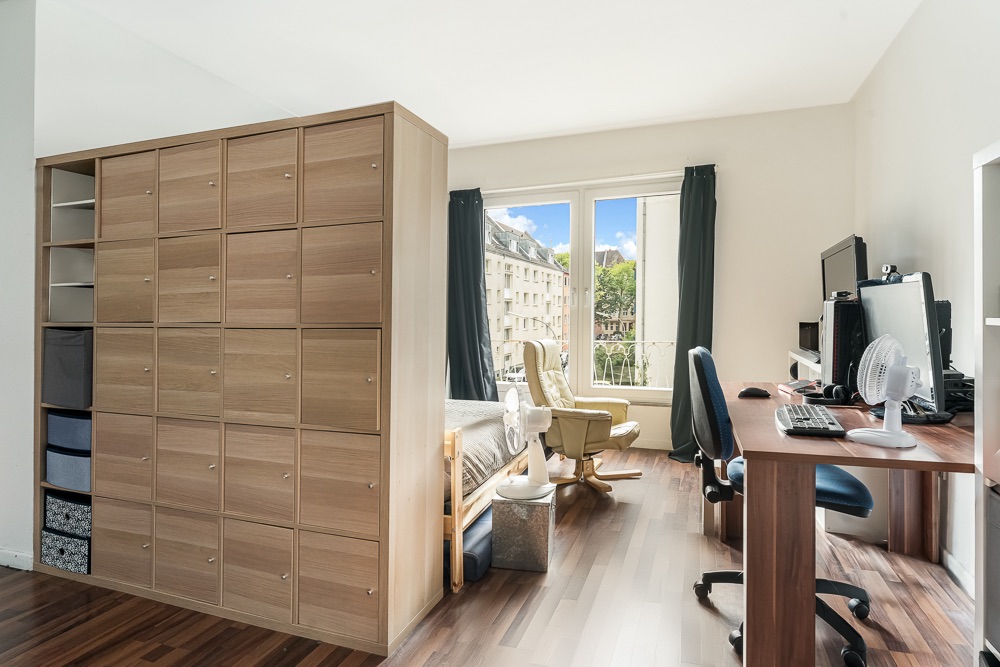 Helles Zimmer mit Schreibtisch, Computer, Regalwand, Bett und großem Fenster.