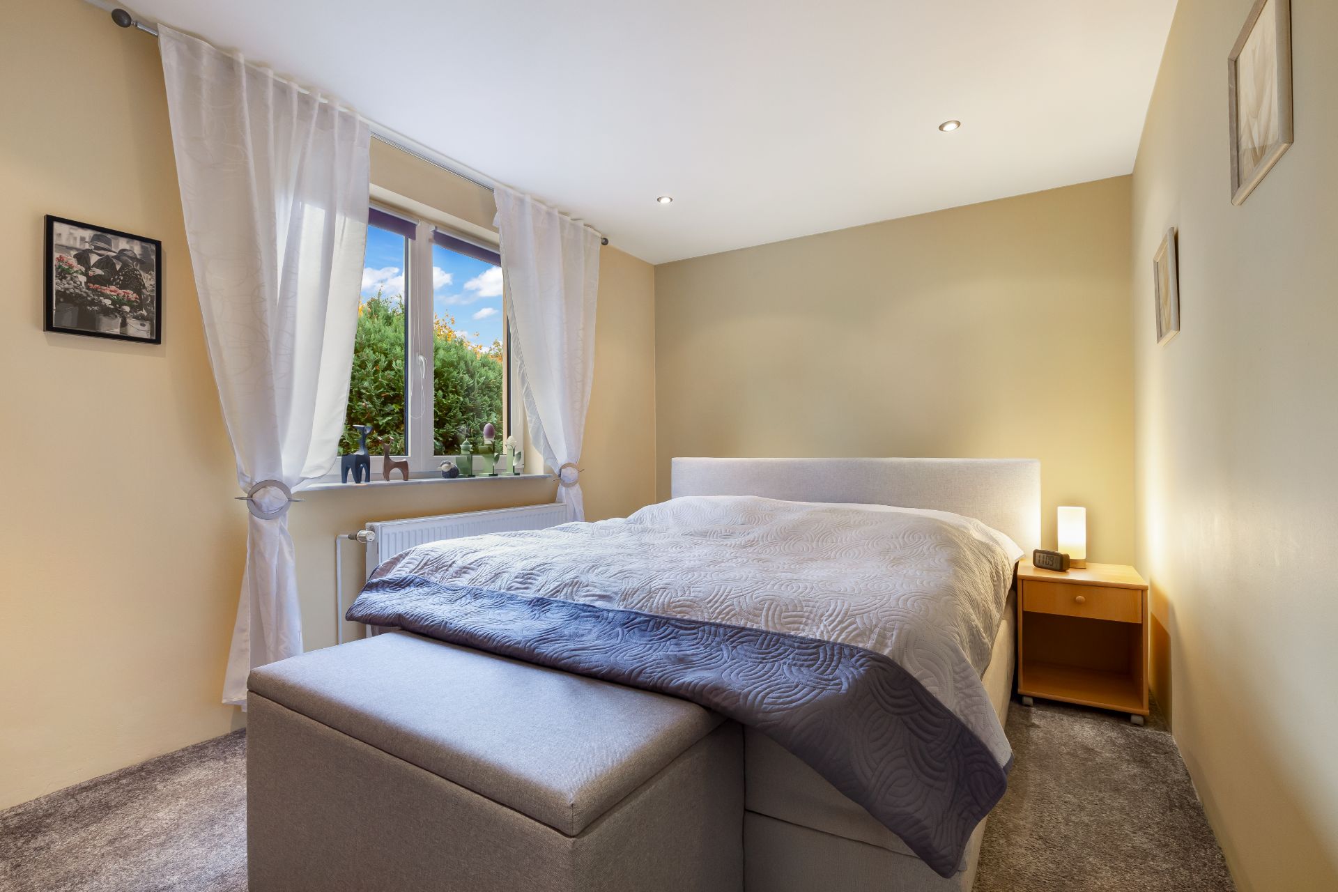 Helles Schlafzimmer mit Doppelbett, Nachttisch, Fenster mit weißen Vorhängen und Teppichboden.