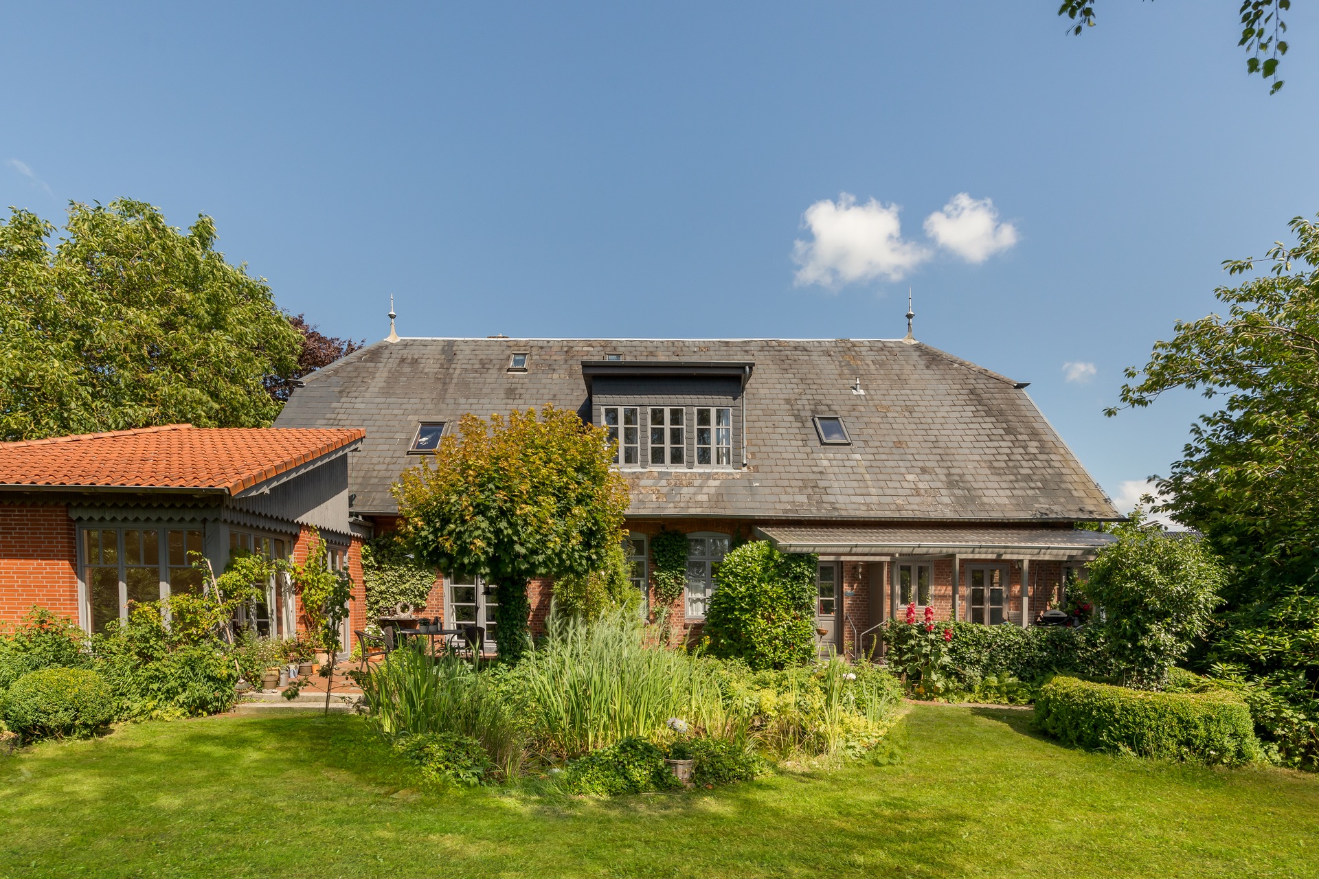 Backsteinwohnhaus mit großem Garten, Teich und Terrasse, umgeben von Bäumen und Blumen.