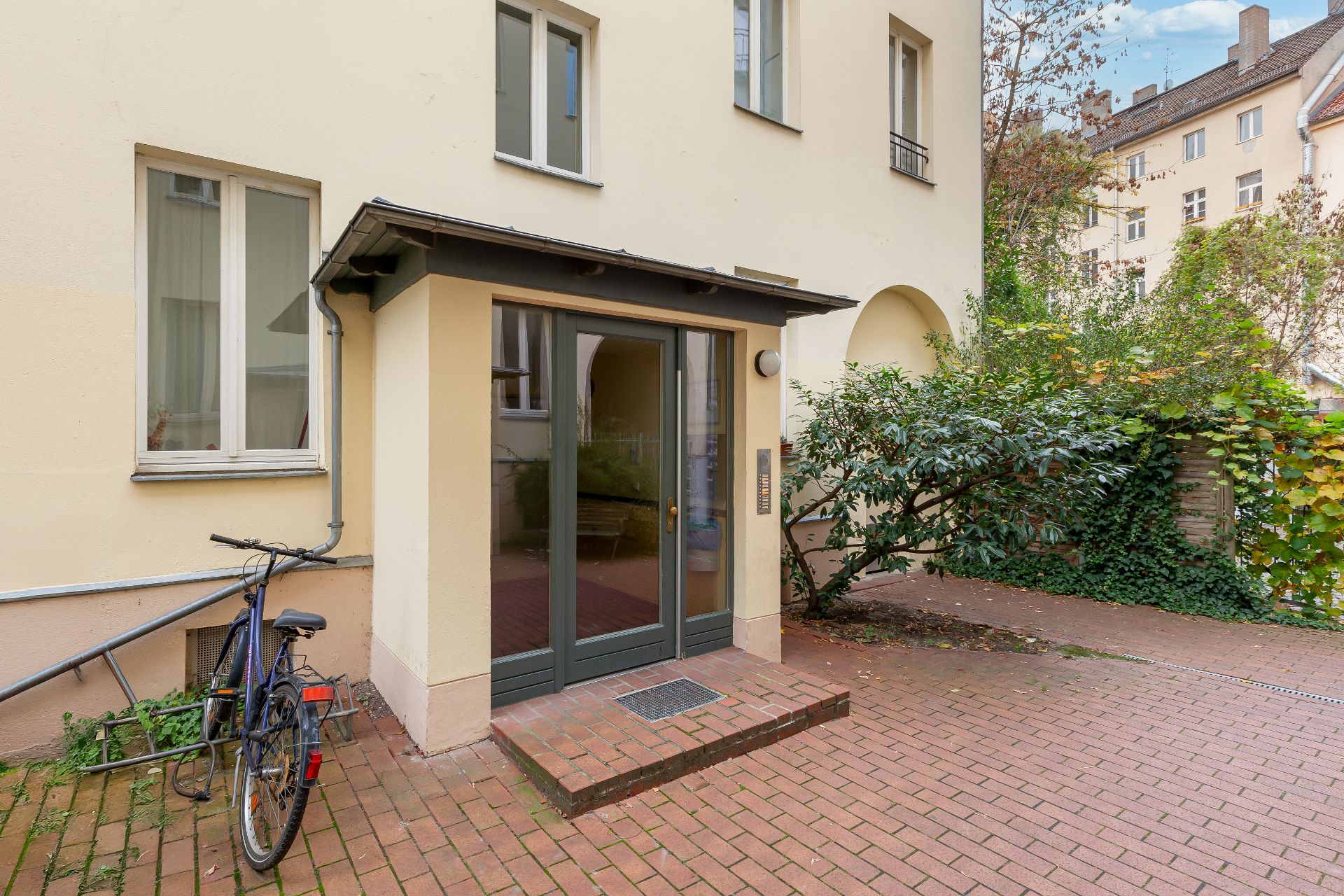 Eingang eines Wohnhauses mit Glasdoppeltür, Fahrrad, Pflanze und gepflastertem Weg.