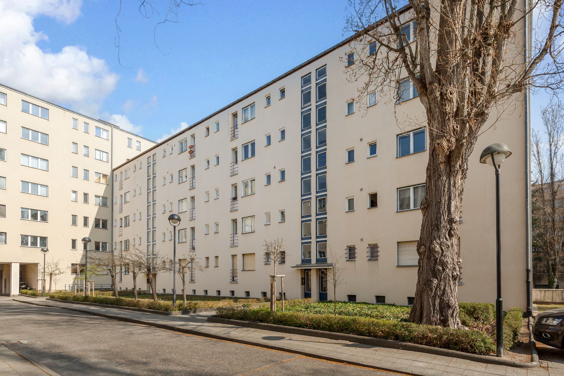 Mehrstöckiges Wohngebäude mit vielen Fenstern, Bäumen und Gehweg im Vordergrund.