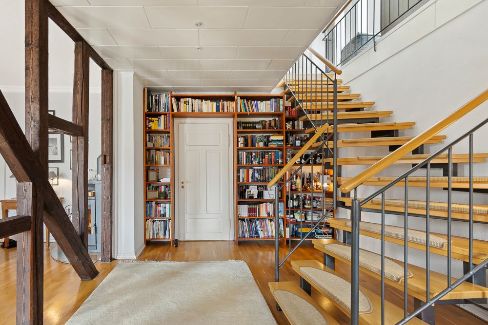 Helles Wohnzimmer mit Bücherregal, weißer Tür und moderner Holztreppe mit Metallgeländer.