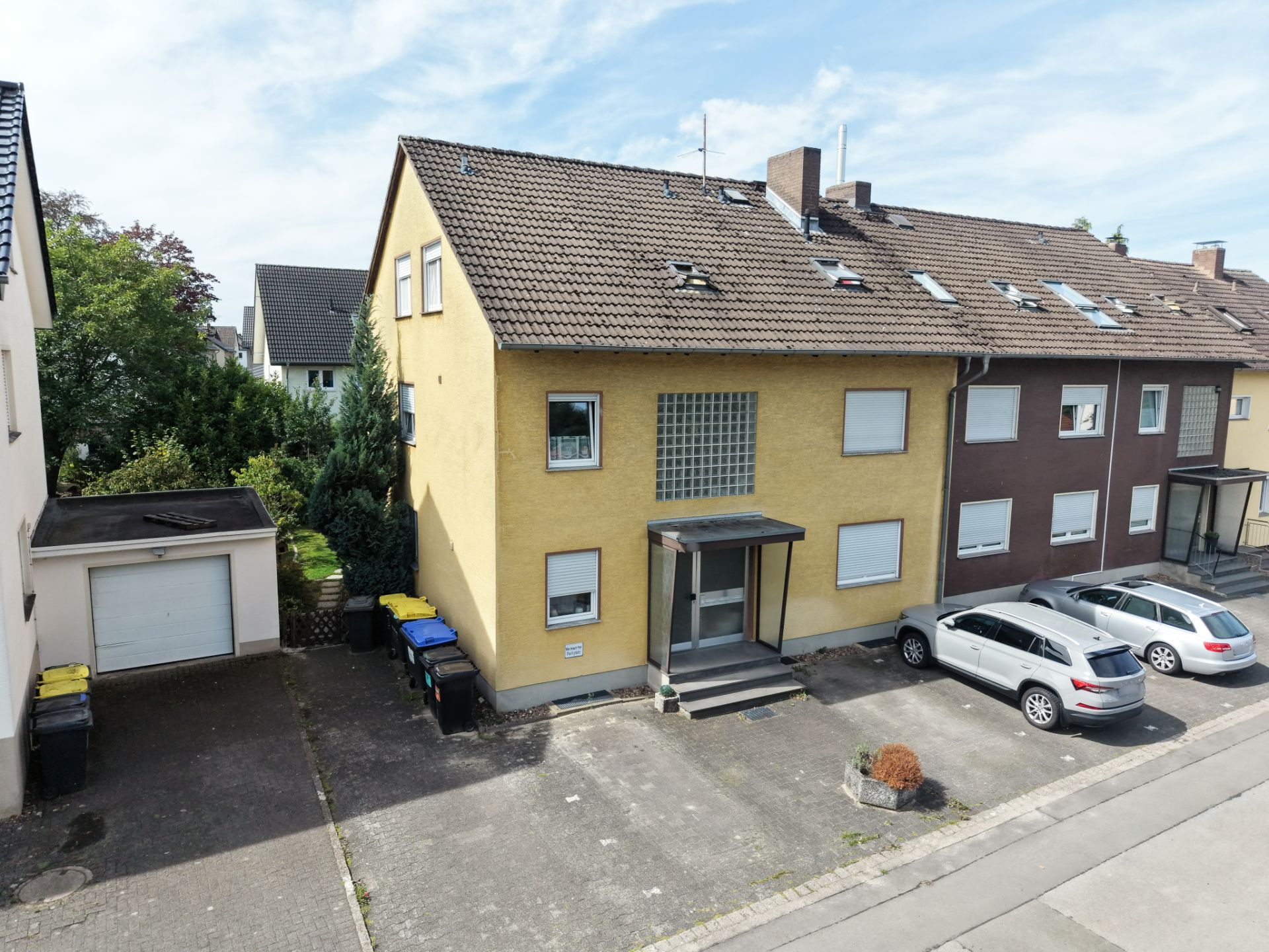 Mehrfamilienhaus mit gelber Fassade, Parkplätzen und angrenzender Garage, aufgenommen bei Tageslicht.