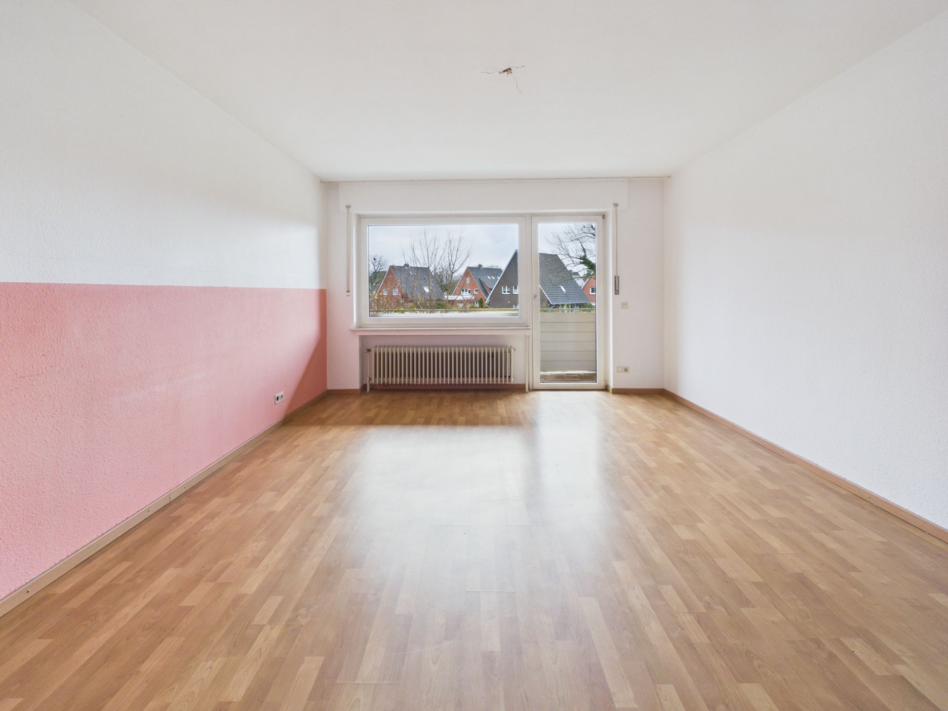 Leeres Zimmer mit Holzboden, rosa-weißer Wand und großem Fenster zur Terrasse.