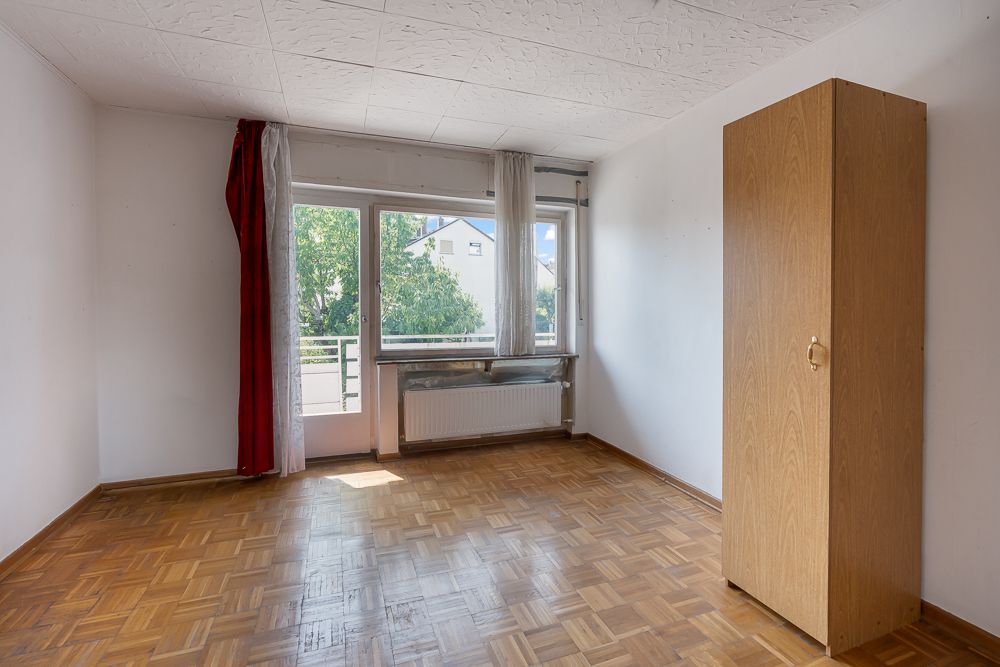 Helles Zimmer mit Parkettboden, Fenster, Vorhängen und einem Holzschrank an der rechten Wand.