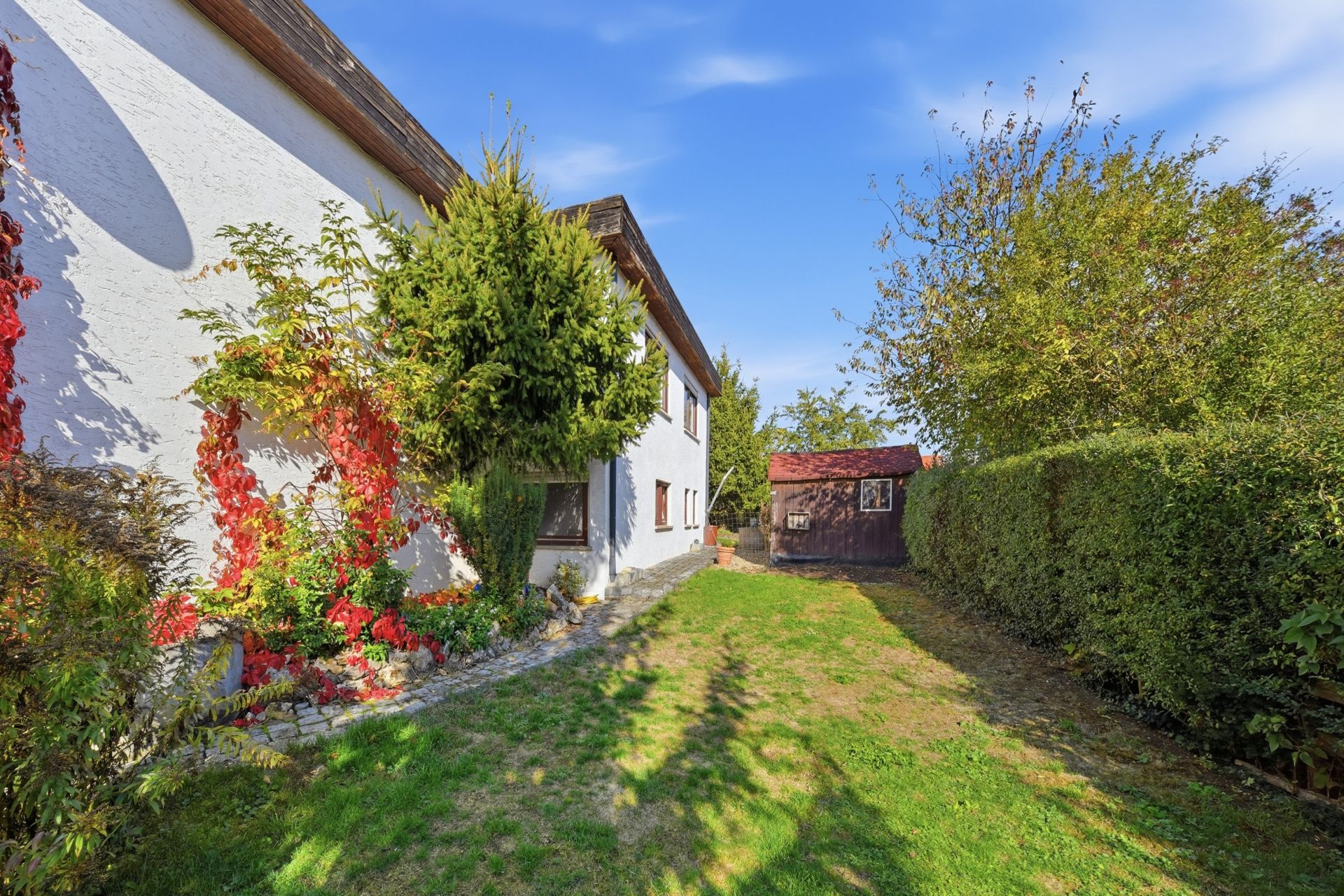 Garten mit Rasen, Sträuchern und einem weißen Haus, im Hintergrund eine kleine Holzhütte.