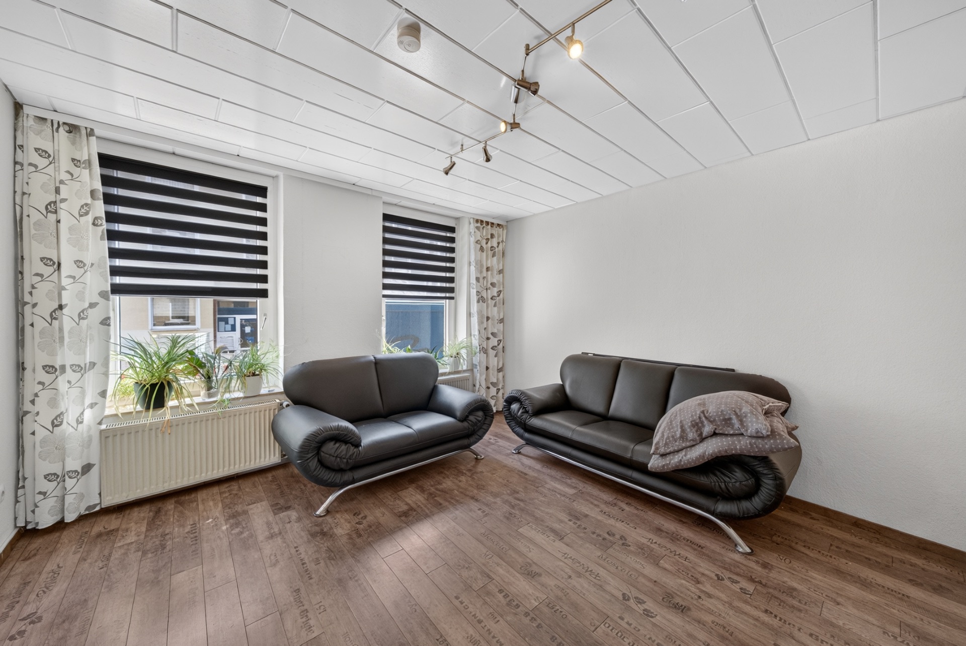Wohnzimmer mit zwei schwarzen Ledersofas, Holzfußboden und zwei Fenstern mit Jalousien.