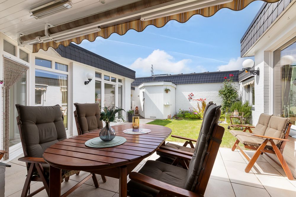 Überdachte Terrasse mit Holztisch und Stühlen, Blick auf einen kleinen Garten mit Rasen und Blumenecke.