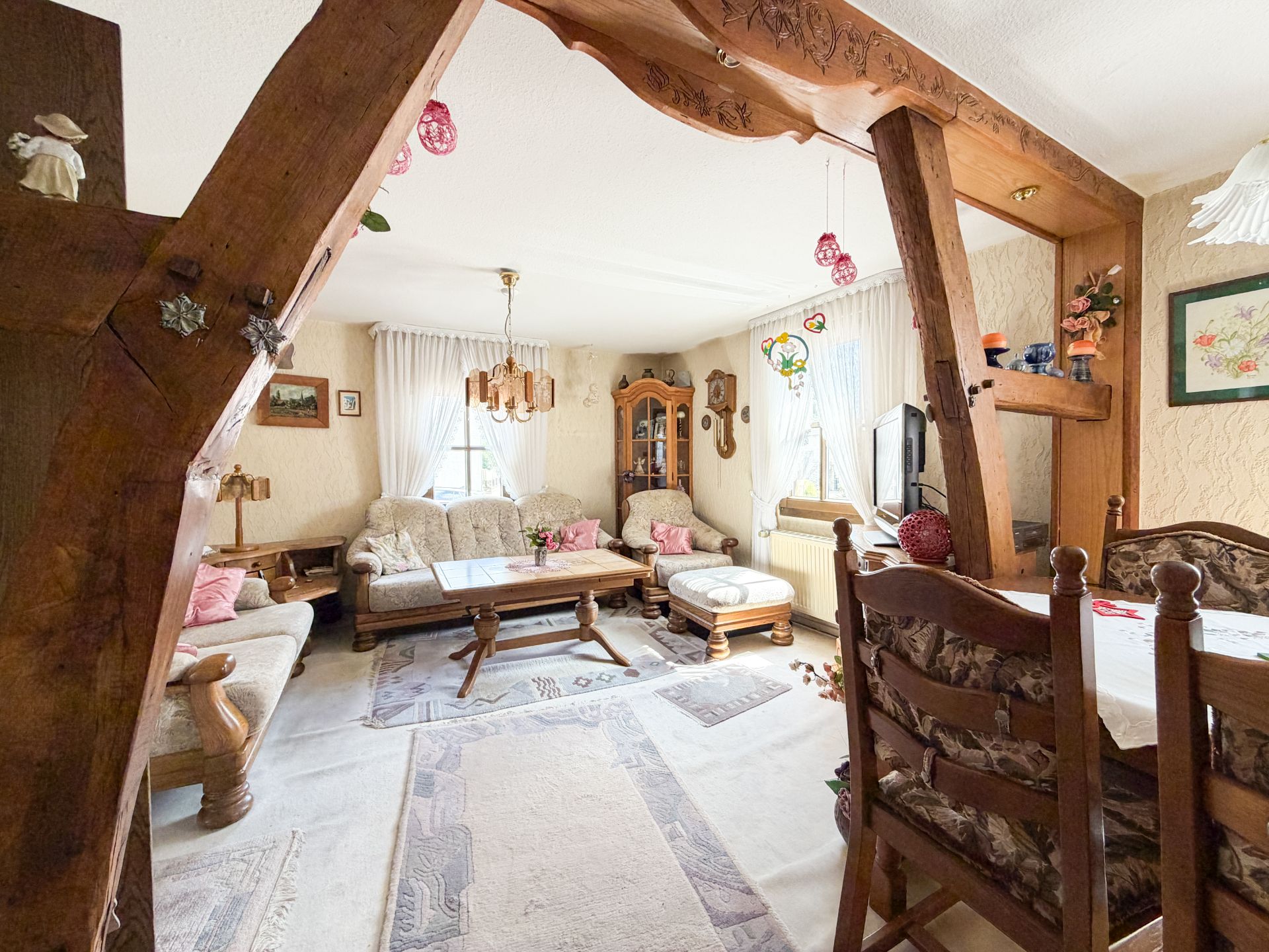 Gemütliches Wohnzimmer mit rustikalem Holzbalken, Sofas, Esstisch und großen Fenstern mit weißen Vorhängen.