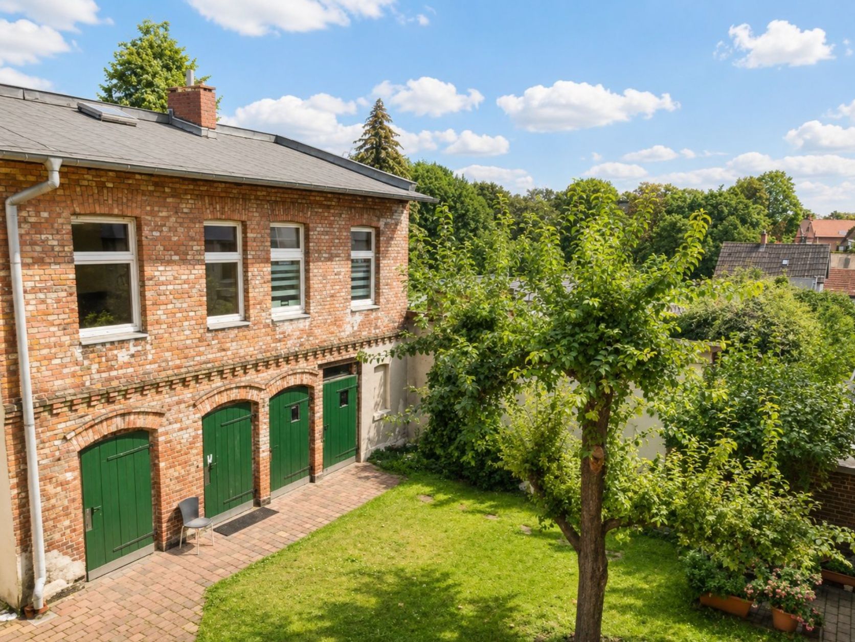 Backsteingebäude mit grünen Türen, Garten und Bäumen an einem sonnigen Tag.